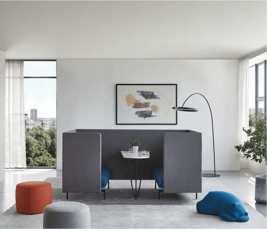 Modern room with gray pod and side partitions, a white table with plants, tall lamp, abstract art, and city view. Orange and gray poufs add color.