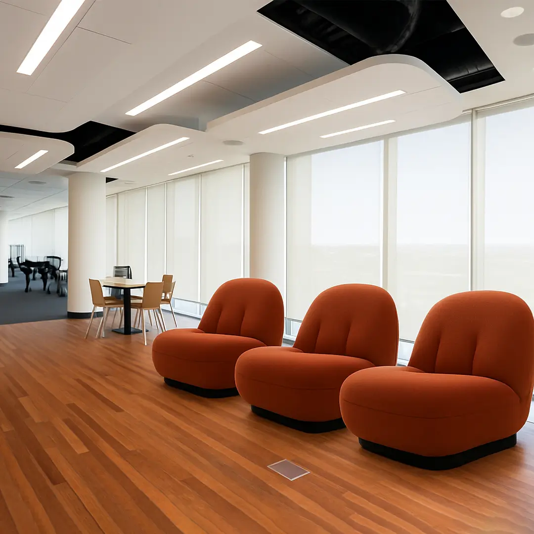 Modern office lounge with three cozy, orange chairs on a wood floor. A round table with four chairs sits by panoramic windows, creating a bright, inviting space.