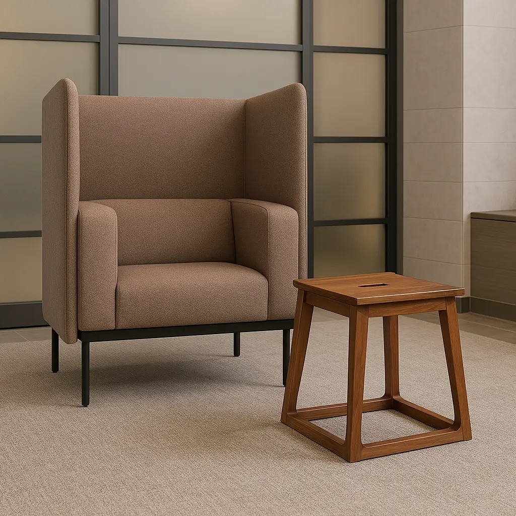 A modern grey armchair with high sides stands beside a small wooden side table on a beige carpet, set against a frosted glass and metal-paneled wall.