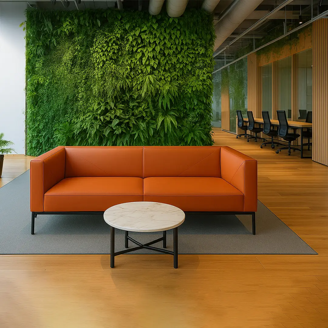 Modern office space with an orange sofa, round marble table, and a lush green vertical plant wall. Open office area with black chairs is visible behind.