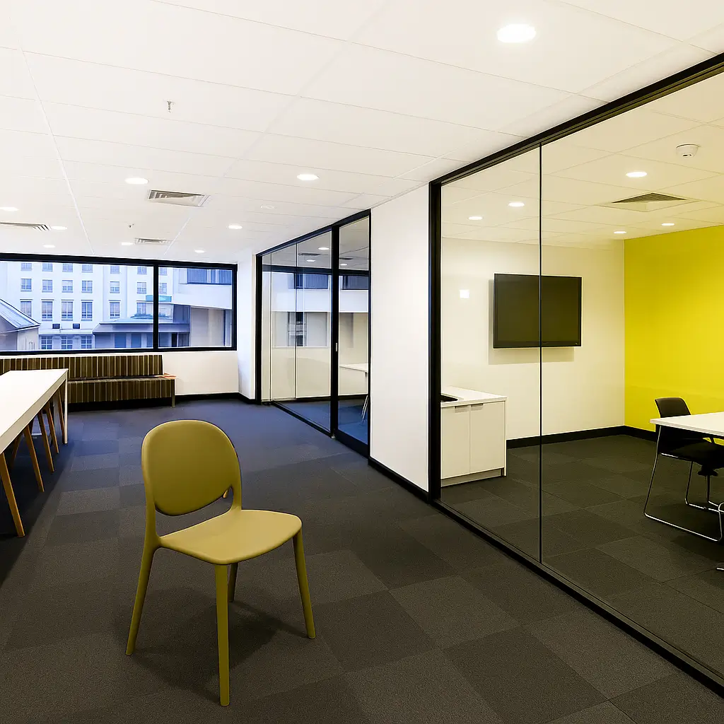 Modern office space with a olive-green chair in the foreground, glass-walled meeting room with a yellow accent wall, and large windows providing natural light.