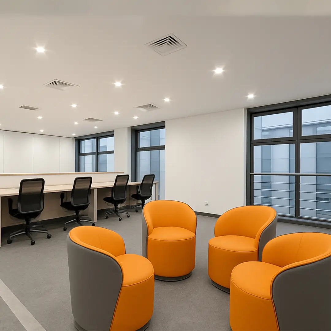 Modern office space with gray carpet, featuring four bright orange chairs in the foreground and a wooden desk with black office chairs in the back.