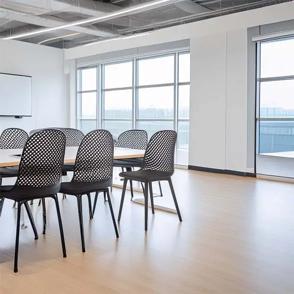 Modern office space with a light wood floor and large windows, featuring a conference table and black perforated chairs. The room is bright and minimalist.