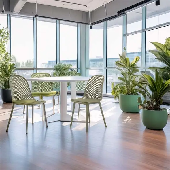 Bright office space with a round white table, four green mesh chairs, and large potted plants by tall windows, creating a fresh, airy atmosphere.