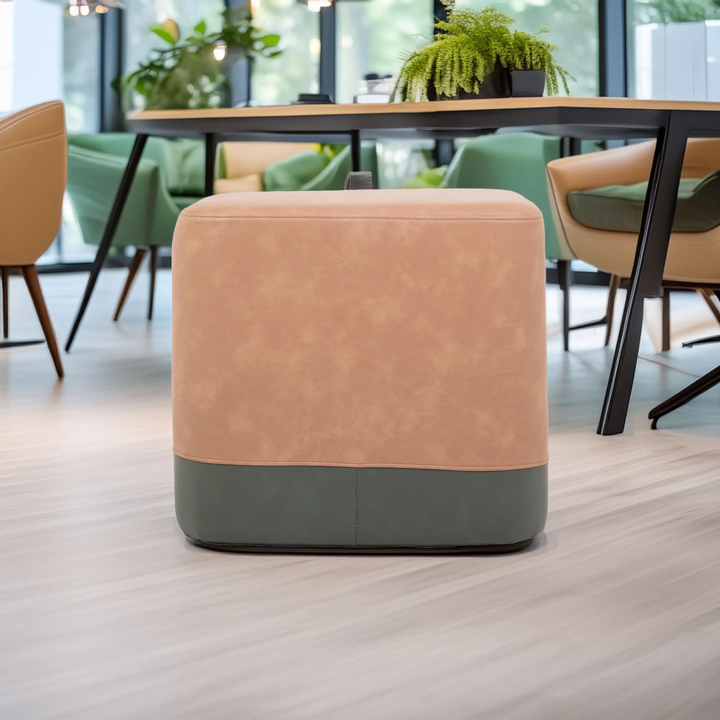 A two-toned ottoman with a tan upper and green base sits on light wood flooring. Background features modern chairs, a table, and lush plants by windows.
