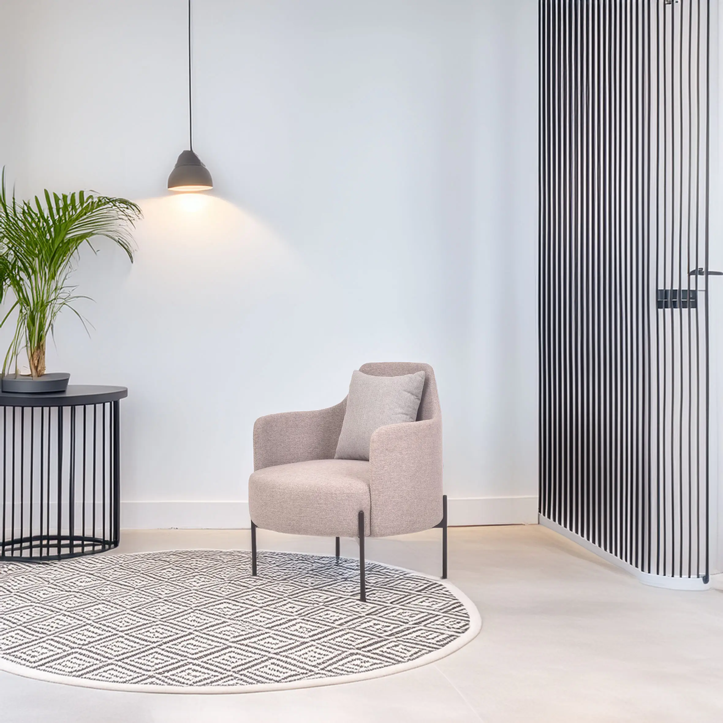 A modern, minimalist room featuring a light grey armchair on a geometric round rug, adjacent to a potted plant and black side table, with a pendant lamp above casting a warm glow.